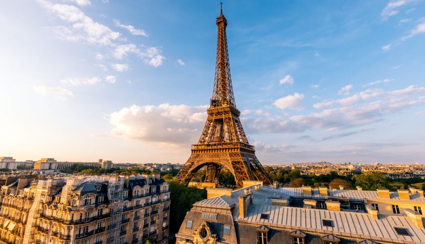 Daytime photo of the Eiffel Tower seen from above, with traditional Parisian rooftops in the foreground. Represents life, travel, and opportunity in France.